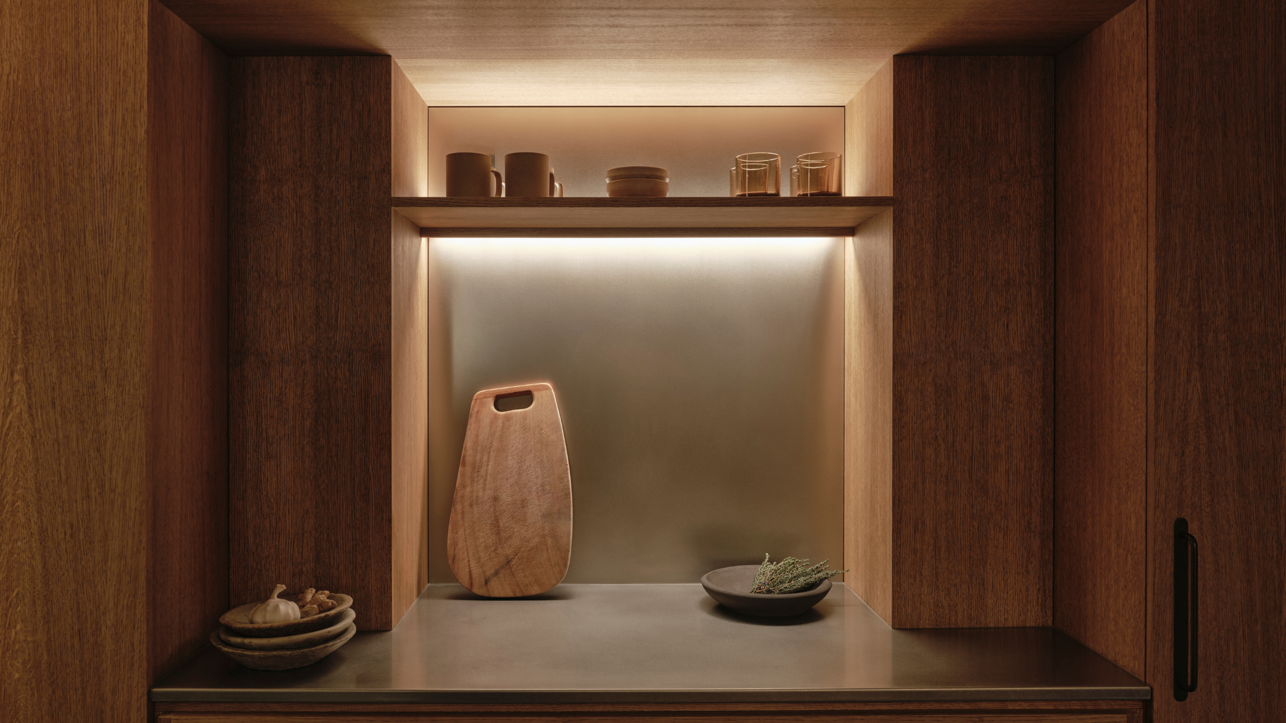 A kitchen in a Japanese home with a backlit shelf, a leaning cutting board, a bowl, and some glasses
