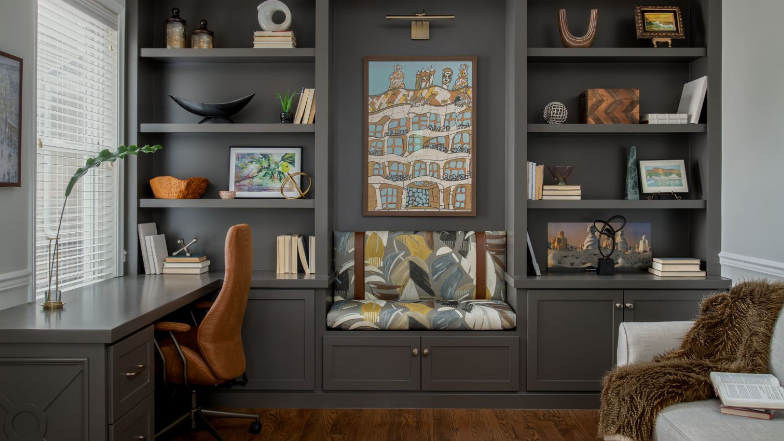 a study room with a gray bookshelf, miscellaneous bookshelf decor, a desk with a brown chair, and a couch with a fuzzy blanket