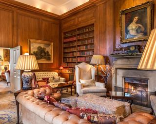 A wood panelled room with Chesterfield leather sofa, mantelpiece, arm chair, floor lamps, book shelves and antique wall art in gold frames