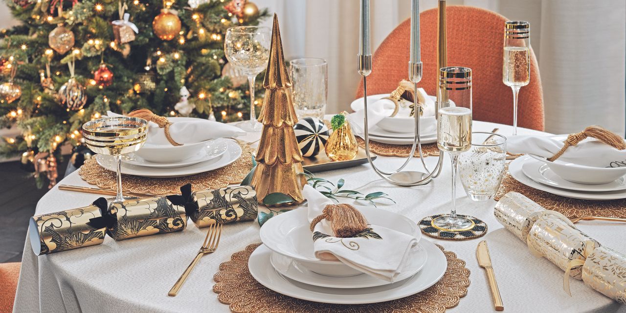 A dining table with a white tablecloth, gold cutlery and candles on top. Decorated for Christmas with gold tree ornaments and Christmas crackers