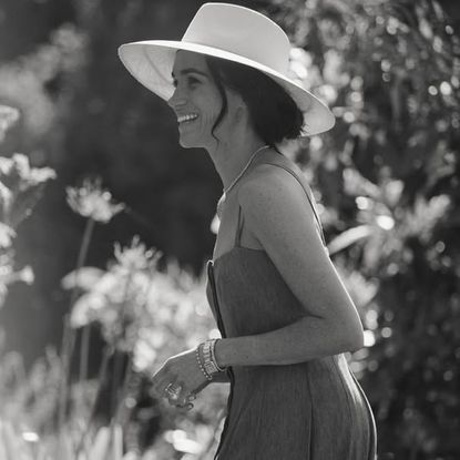 Meghan Markle wearing a sleeveless dress and sun hat smiling and turning to the side