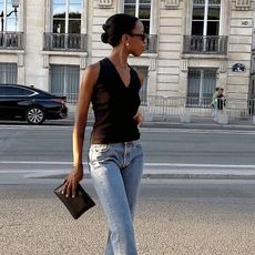 British influencer Sylvie Mus poses on a sidewalk in the 1st arrondissement in Paris wearing gold hoop earrings, a v-neck black sleeveless top, black clutch, and straight-leg jeans
