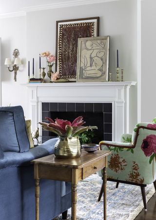A fireplace with paintings on the mantelpiece and a floral chair.