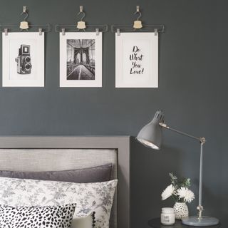 Dark grey bedroom wall with prints hanging from clothes hangers. Below it is a bed and bedside table with a lamp on it