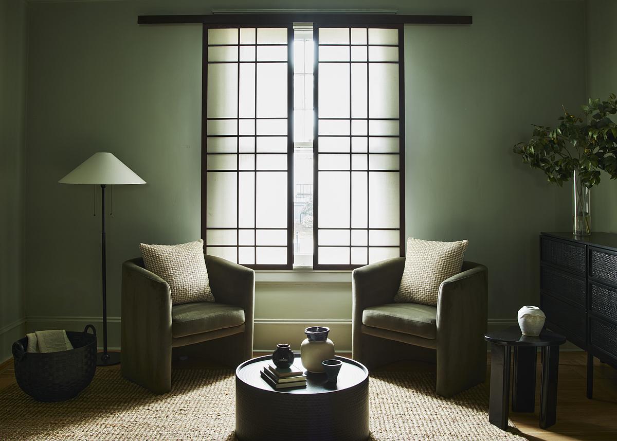 a sage green room with thick carpet, two green armchairs with a cushion, a floor lamp, basket with blanket, round coffee table with vases styled on it, a console table with vase of branches, and a side table