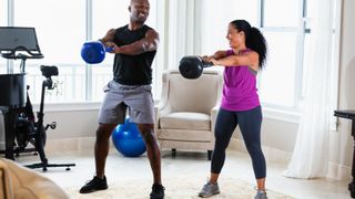 A man and woman perform kettlebell swings in a living room. They are both stood with legs slightly wider than hip-width apart, with their arms straight in front of them holding the kettlebell as it swings out in front of them. Behind them we see an arm chair and an exercise bike.