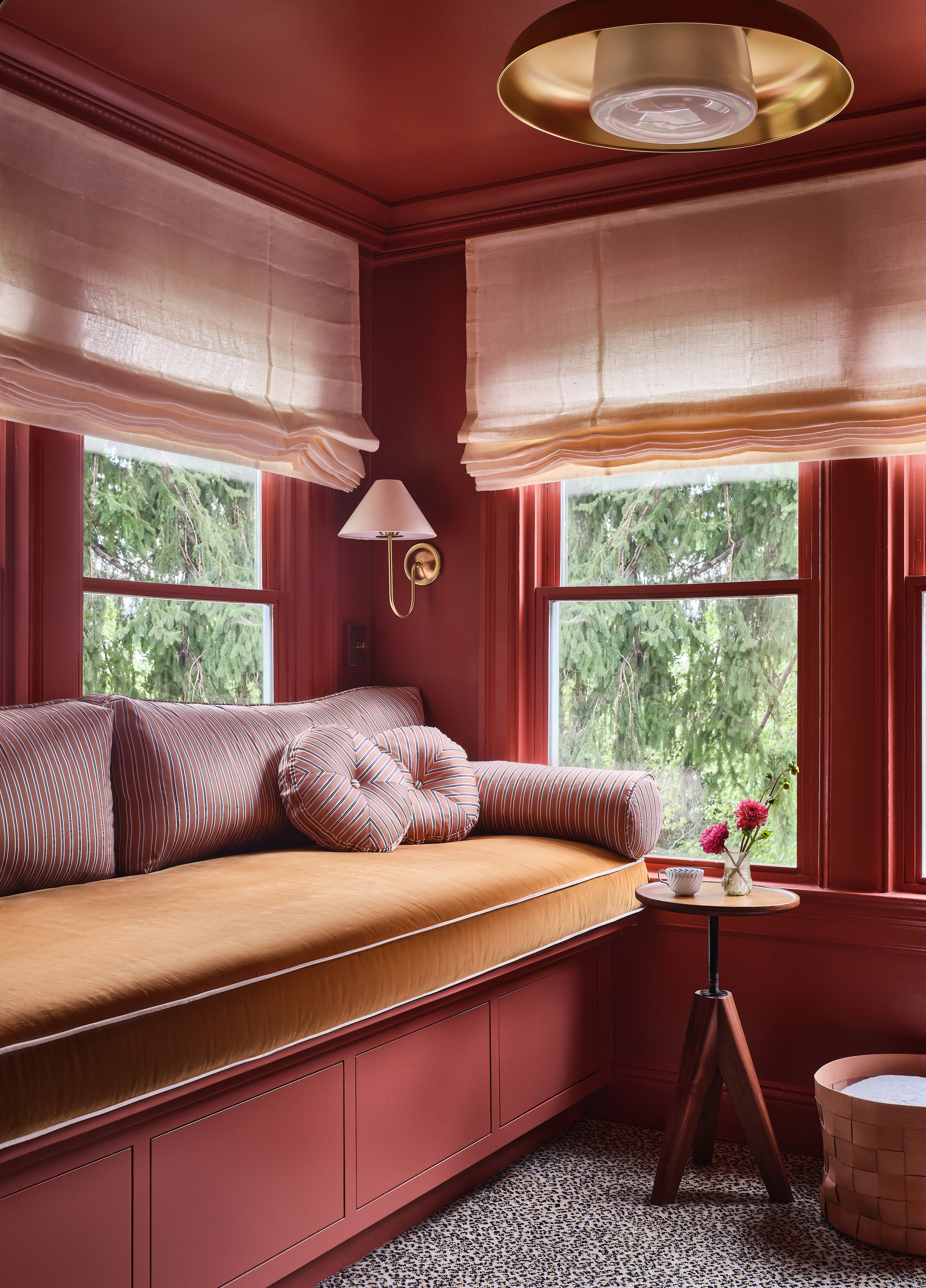 Image of the corner of a lounge room. The walls and ceiling are color drenched in a rich coral red. There is a built-in bench seat that has a golden/ochre yellow cushion and pearl-colored bolster pillows. There is a gold light fixture on the ceiling and a gold sconce light on the wall.
