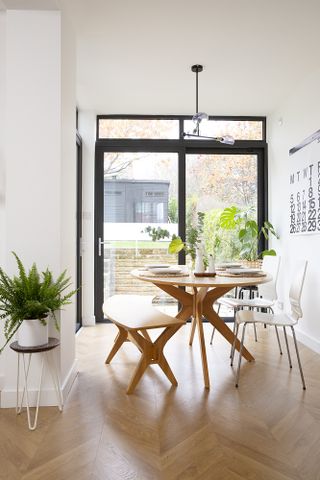 Dining area of an open-plan kitchen in Maria's home with wooden table and bench, white chairs, and black framed sliding doors