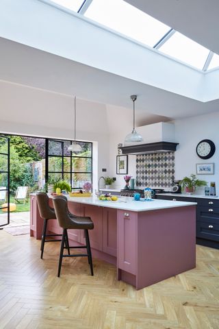 A kitchen with pink kitchen island, pale wooden Herringbone flooring and Harlequin tiled backsplash