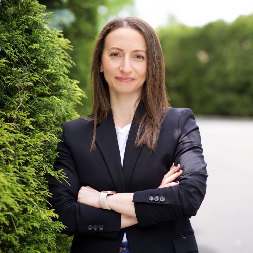 Headshot of Polina Shkadron stood with arms crossed next to foliage.