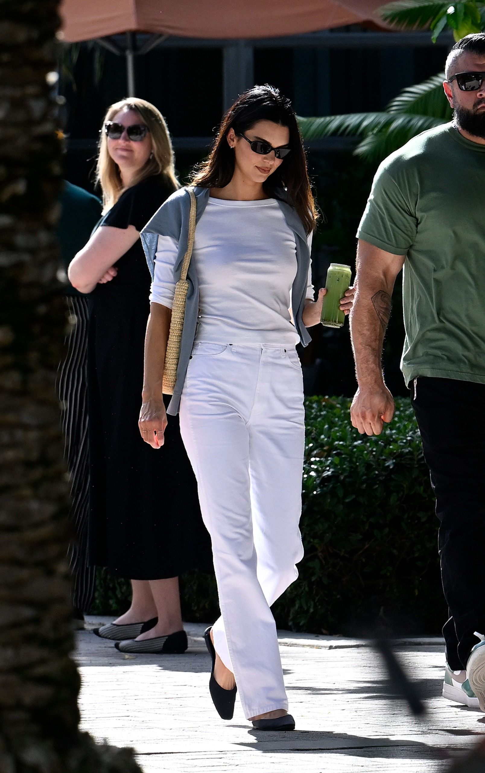 Kendall Jenner walks outside wearing white straight-leg trousers with a white boat-neck t-shirt and black slipper shoes.