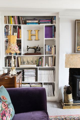 White bookshelf filled with books and trinkets behind purple sofa with blue embroidered cushions