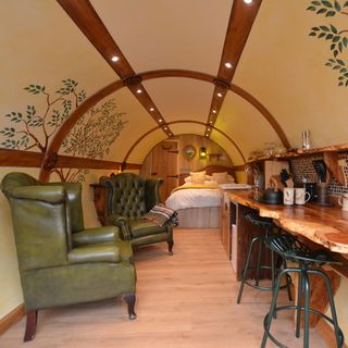 the inside of a hobbit house showing it's curved roof and walls with a tree design painted on it, a small open plan space with the bed at the back, and then towards the front two green leather armchairs on the left and a long wooden bench kitchen on the right
