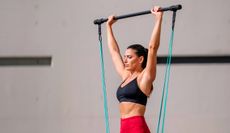 A woman in sportswear uses a Pilates bar. She holds the bar above her head with her hands wider than shoulder-width apart. Her arms are extended. Blue resistance bands stretch from each end of the bar towards her feet.