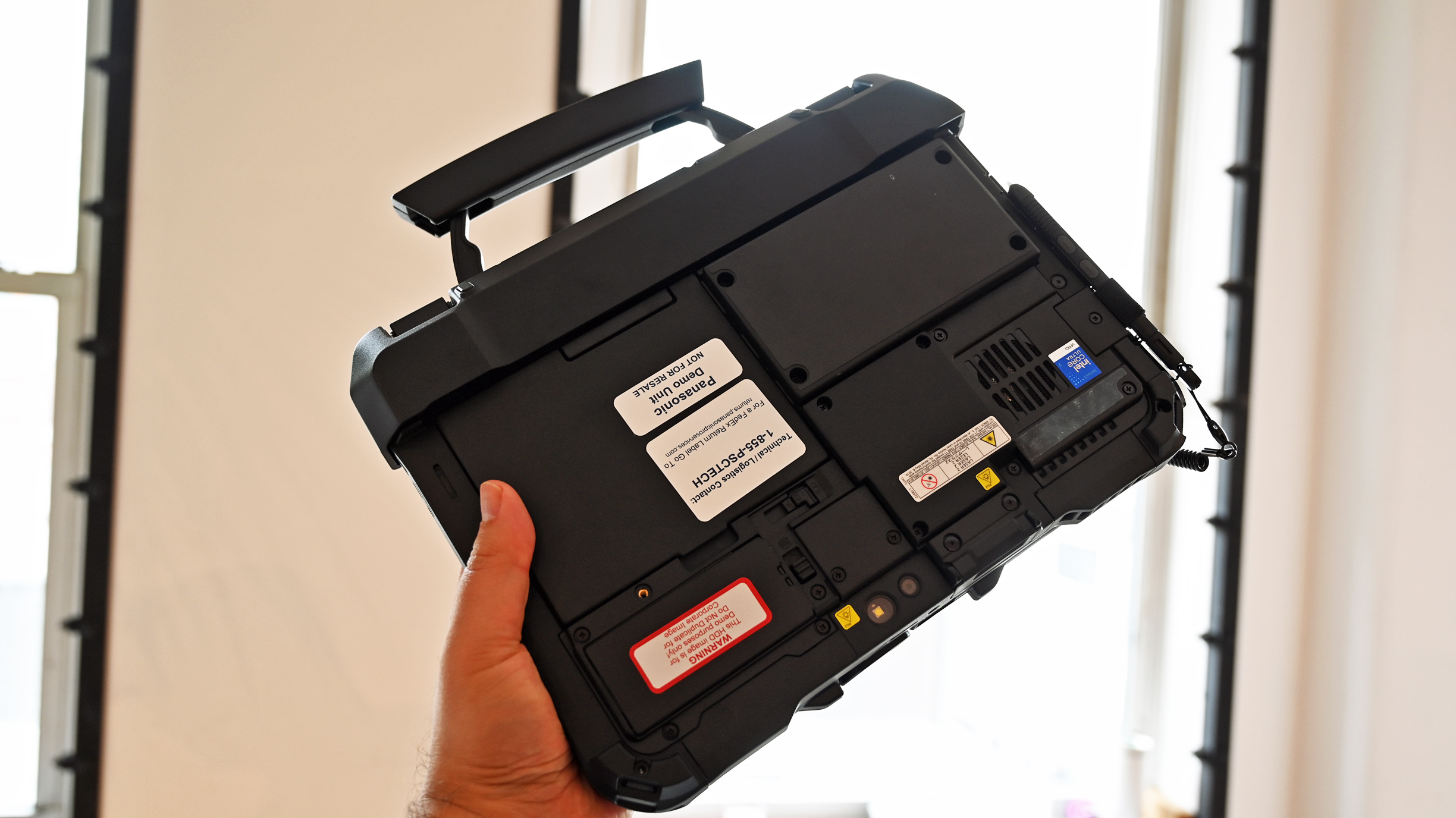 A person holding a rugged Panasonic TOUGHBOOK G3 by its top carrying handle, showing the back chassis and component labels.