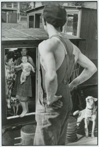 A woman holding a baby stands next to an older woman in the doorway of a barge, as seen from over the shoulder of a man facing them.