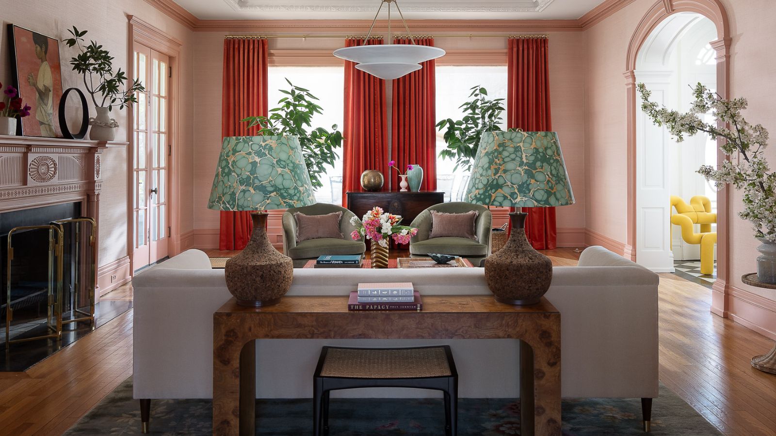 pink open living room with red curtains at the window, a white sofa with a burl wood console table behind it with two lamps with green marbled shades
