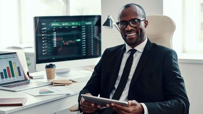 A smiling fund manager sits in front of multiple computers showing investment information