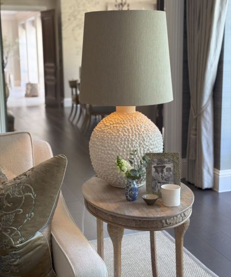 Close up of large lamp, textured round base with green lampshade, sat on top of round wooden side table with framed picture and ornaments, cream sofa to the left