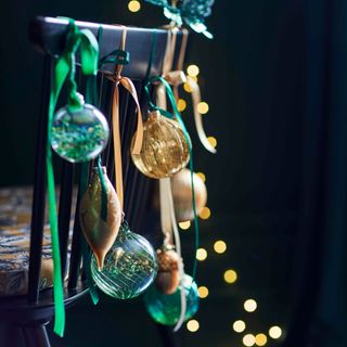 green and gold baubles and ribbon hanging off a black dining chair, with lights glowing in the background