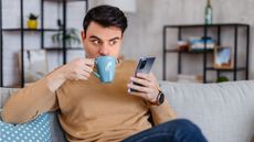 A man drinks coffee while he talks on speakerphone while sitting on his sofa.