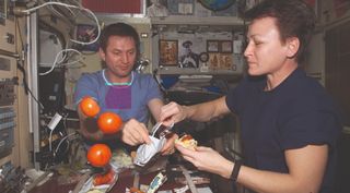 Two people attempt to prepare food in zero gravity. Some food appears to be "floating" alongside them.