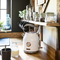 A white kettle on a wooden countertop. Next to it is a black mug and a small bottle of milk.