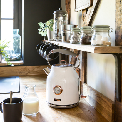 A white kettle on a wooden countertop. Next to it is a black mug and a small bottle of milk.