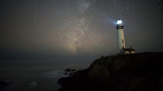 A line of light stretches across a star-studded sky next to a little white lighthouse perched on the rocks. 