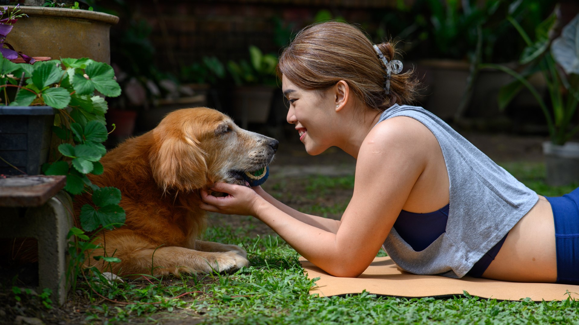 a smiling woman lies on a yoga mat and pets her senior golden retriever in a yard