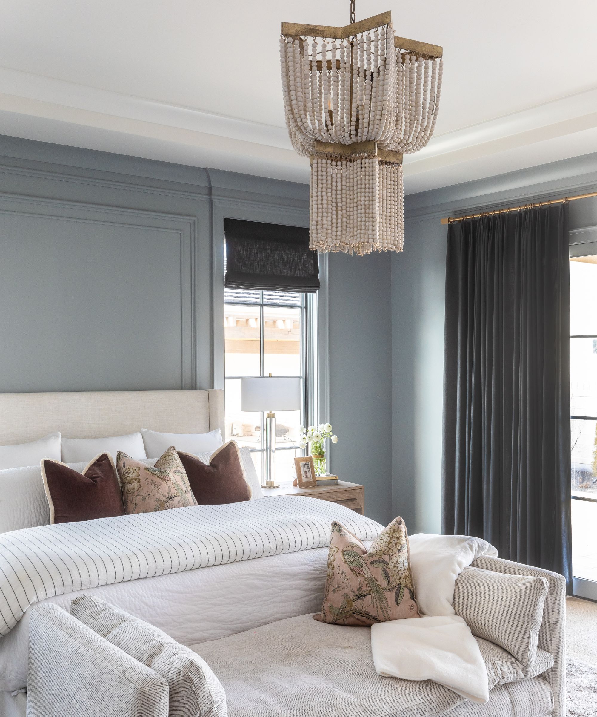 Bedroom with Boothbay Gray painted walls, glass doors with drapes, an upholstered cream bed with brown throw pillows and white bedding, a linen daybed at the foot of the bed, and a chandelier above.