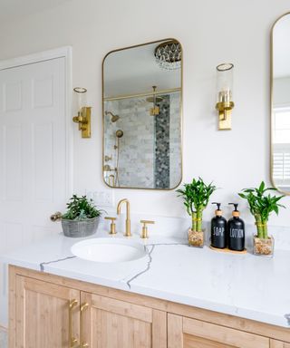 A bathroom with a curved gold mirror, a sink unit with wooden cabinets, a marble countertop, and gold taps and plants and soap dispensers on it