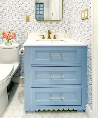 A half bath with a blue scalloped vanity with a white counter and gold faucets, blue and white patterned wallpaper on the walls and a gold mirror, and a white toilet to the left with pink roses on top
