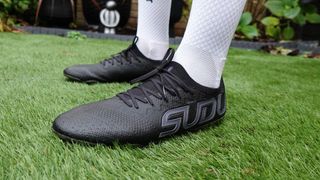 A person wearing white socks is standing in a pair of black football boots with 'SUDU' displayed on the side of one, standing on green artificial grass. SUDU SFS FG 01+ Pro