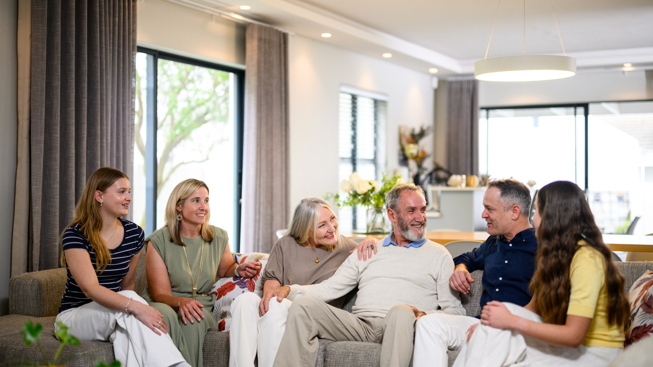 A group of family or friends of multiple generations sits on a couch, talking.