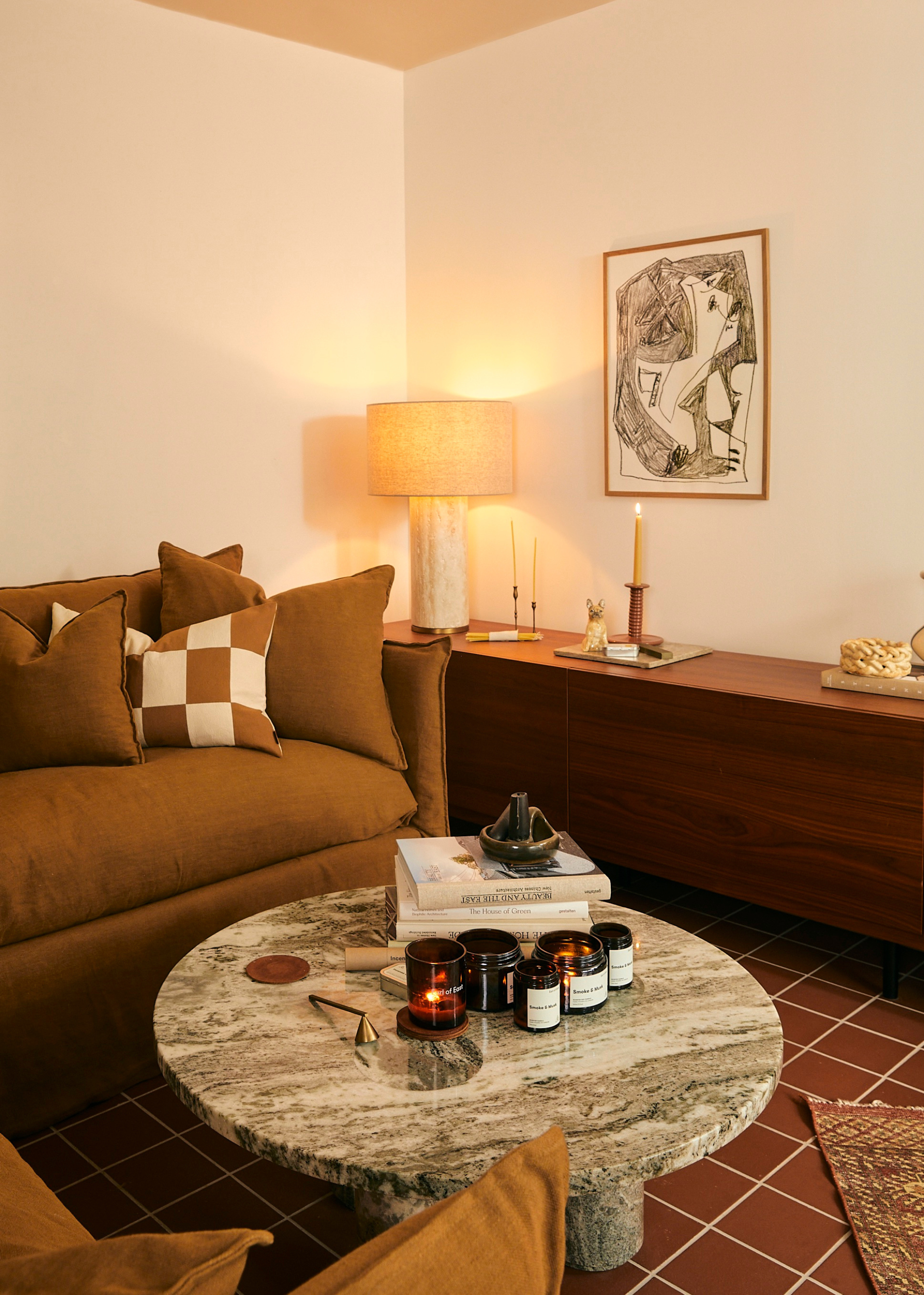 A living room corner with a brown couch, a green marble table with a cluster of candles, a snuffer, and a stack of books by a sideboard with a table lamp, and a taper