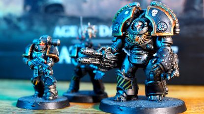 A Saturnine Terminator and two MKII Crusader pattern Space Marines on a wooden surface in front of the Saturnine box