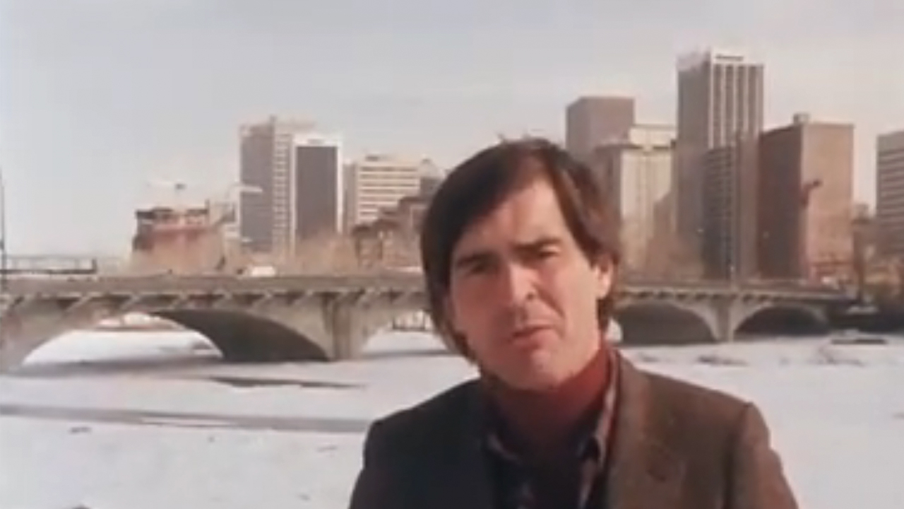 A reporter on the banks of the Bow river, in Calgary, with the city and a bridge in the background
