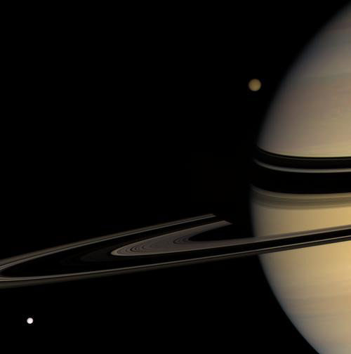 Titan emerges from behind Saturn while Tethys streaks into view in this colorful scene on March 24, 2008. Titan is 5,150 kilometers (3,200 miles) wide; Tethys is 1,071 kilometers (665 miles) wide. Saturn&#039;s shadow darkens the far arm of the rings near the 