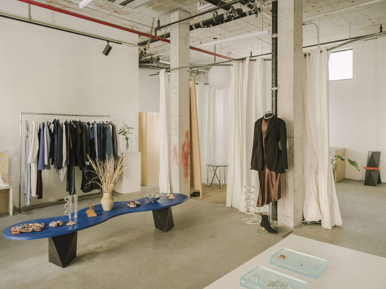 A whimsically furnished clothing store features a wavy blue and black coffee table covered in sculptural creations and clothing hanging in an industrial-style, cream-shaded space.