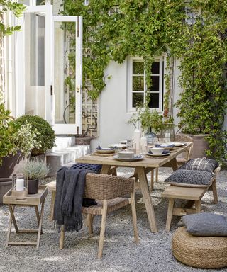 How to plan an outdoor kitchen in a gravel patio with natural wooden table, bench seating, wicker chairs and footstools, surrounded by climbing and potted plants and gray textiles.