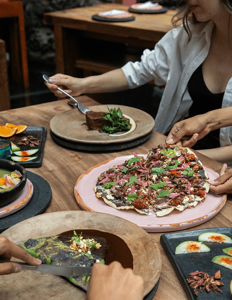 A table captured from above shows several people as they prep their tacos and quesadillas on a wooden table decorated with flat ceramic tableware.