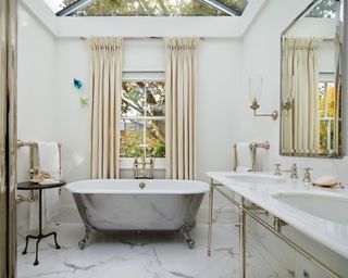 An example of how to make a small bathroom look bigger showing a rolltop bath beneath a window with curtains, a marble floor and double vanity unit