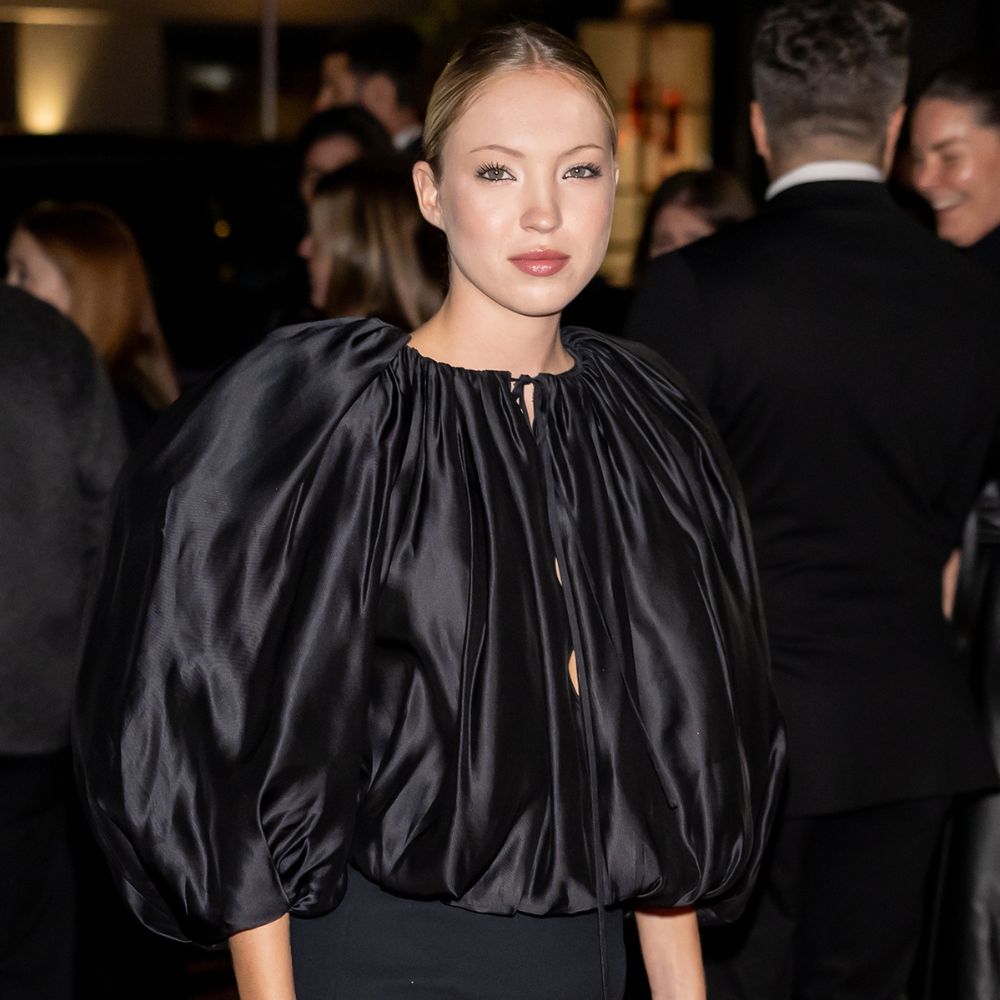 Lila Moss walks a red carpet wearing a black satin balloon blouse with a black miniskirt and black pointed-toe heels.