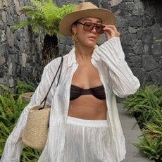 Woman wears straw hat, sunglasses, crinkle shirt, brown bikini top, white shorts and basket bag