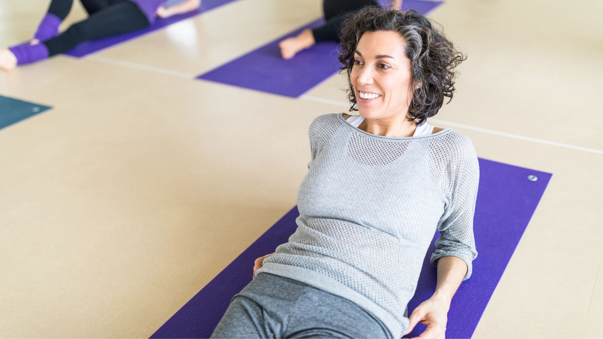 Woman lying down on yoga mat in class, wearing activewear and smiling, in set-up for the reverse plank
