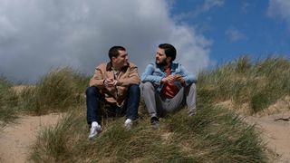 Sam and Asif sit on a sandy dune at the beach talking with blue sky behind them