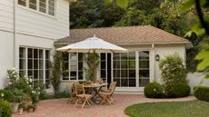 the exterior of a neutral clad home with a brick patio and curved lawn with wooden patio furniture and terracotta pots with flowers