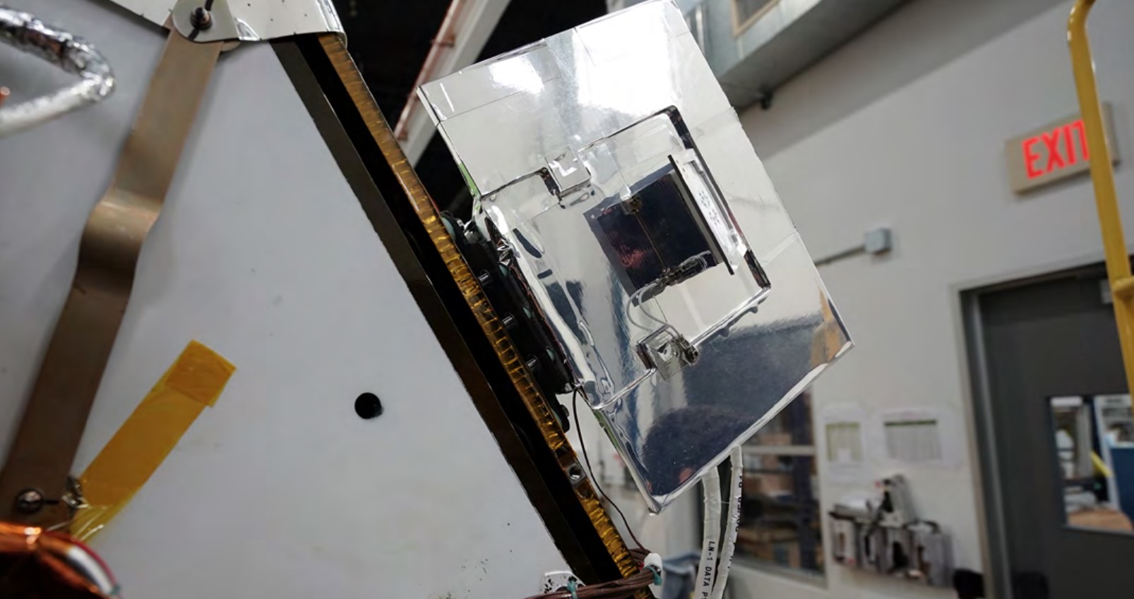a small box wrapped in metallic foil on the side of a tall rectangular spacecraft on four legs