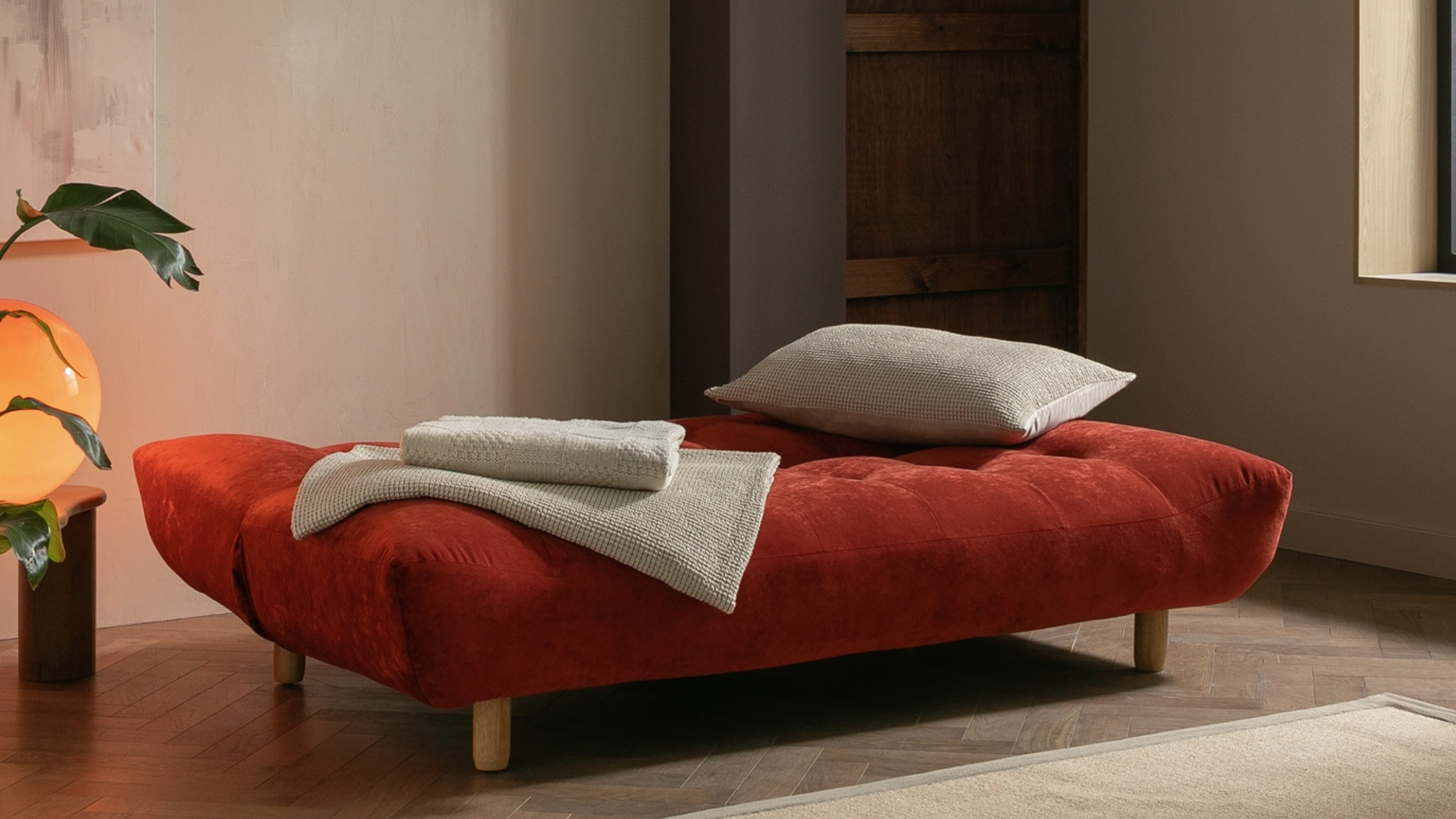 Habitat Kota 3-Seater Sofa Bed in Burnt Orange set up as a bed with a pillow and bedding folded on top ready to be made in a minimalist space with layered lighting and a plant